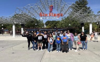 Group of iQ Academy students, teachers, and families posing together in front of the Six Flags Magic Mountain entrance on a sunny day.