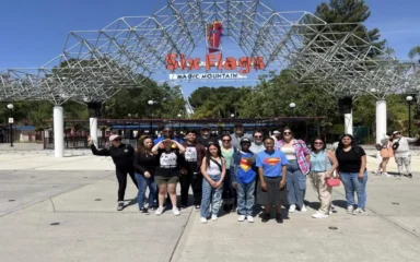 Group of iQ Academy students, teachers, and families posing together in front of the Six Flags Magic Mountain entrance on a sunny day.