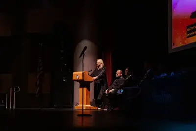 A woman stands at a podium on a dimly lit stage, addressing an audience. Several seated people are partially visible. The atmosphere is formal and focused.