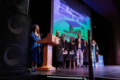 Graduation ceremony stage with a student in a blue cap and gown at a podium. Seven individuals in academic attire stand smiling. Screen reads, "IQ Academy Class of 2023."