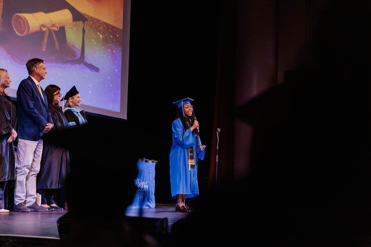 Graduation ceremony on stage; a graduate in a blue gown speaks into a microphone. Four faculty members stand to the left, with a diploma image projected behind.