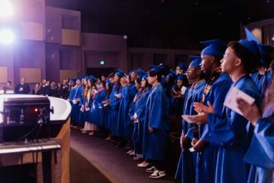 A group of graduates in blue gowns stand in a dimly lit auditorium. They face the front, some clapping. A bright light shines on the left, creating a celebratory atmosphere.