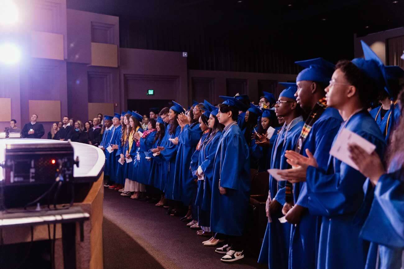 A group of graduates in blue gowns stand in a dimly lit auditorium. They face the front, some clapping. A bright light shines on the left, creating a celebratory atmosphere.