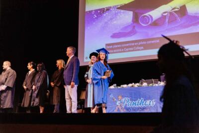 Graduate in blue gown holds diploma on stage during a ceremony. A screen shows a congratulatory image and text. People in robes stand in the background.