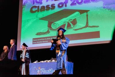 A graduate in a blue cap and gown, smiling and holding a diploma. Behind, a presentation reads "Class of 2025" with a projected image of a cap and diploma.