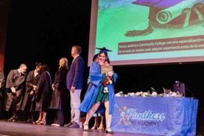 Graduate in blue cap and gown smiles while walking across a stage, holding a diploma. People in gowns stand in line. A decorated table and screen are in the background.