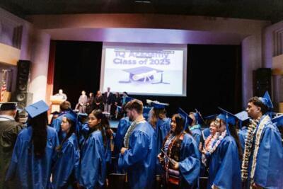 Graduates in blue gowns and caps face a stage with faculty members. A large screen reads "IQ Academy Class of 2025," creating a celebratory atmosphere.