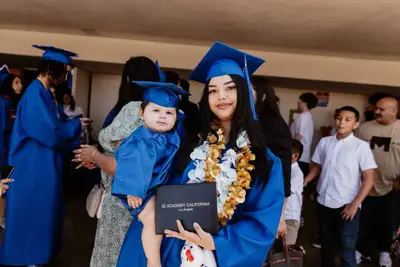 Young woman in a blue graduation gown holds a baby in a matching cap and gown, smiling proudly with a diploma. Celebratory atmosphere with crowd.