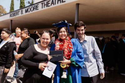 A joyful graduate in a blue cap and gown holds a diploma, surrounded by smiling supporters outside the Robert B. Moore Theatre.