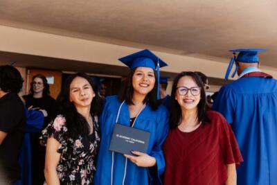 Graduate in blue cap and gown holds diploma, smiling between two women, one in a floral dress, the other in red. Background features others in gowns.