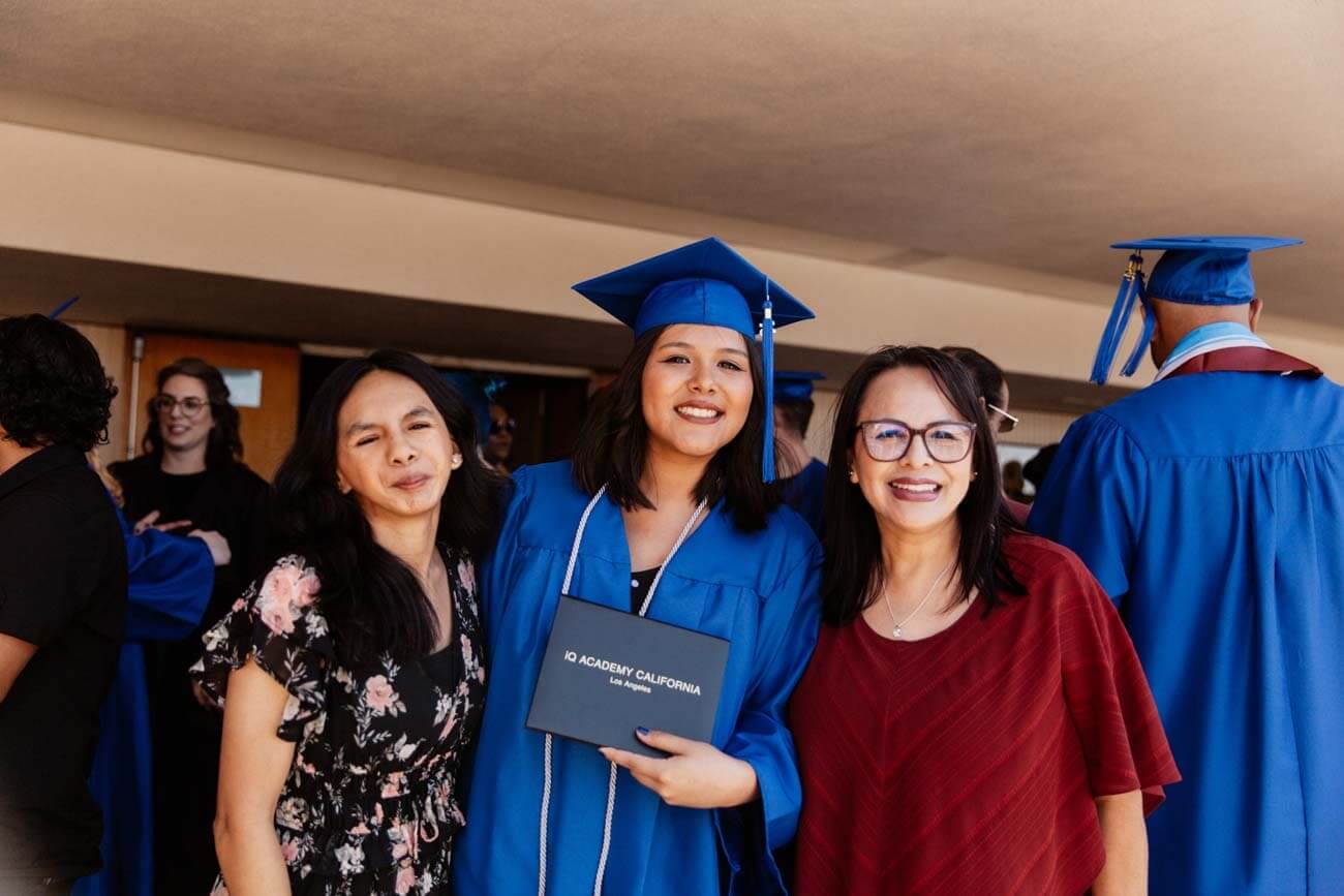 Graduate in blue cap and gown holds diploma, smiling between two women, one in a floral dress, the other in red. Background features others in gowns.