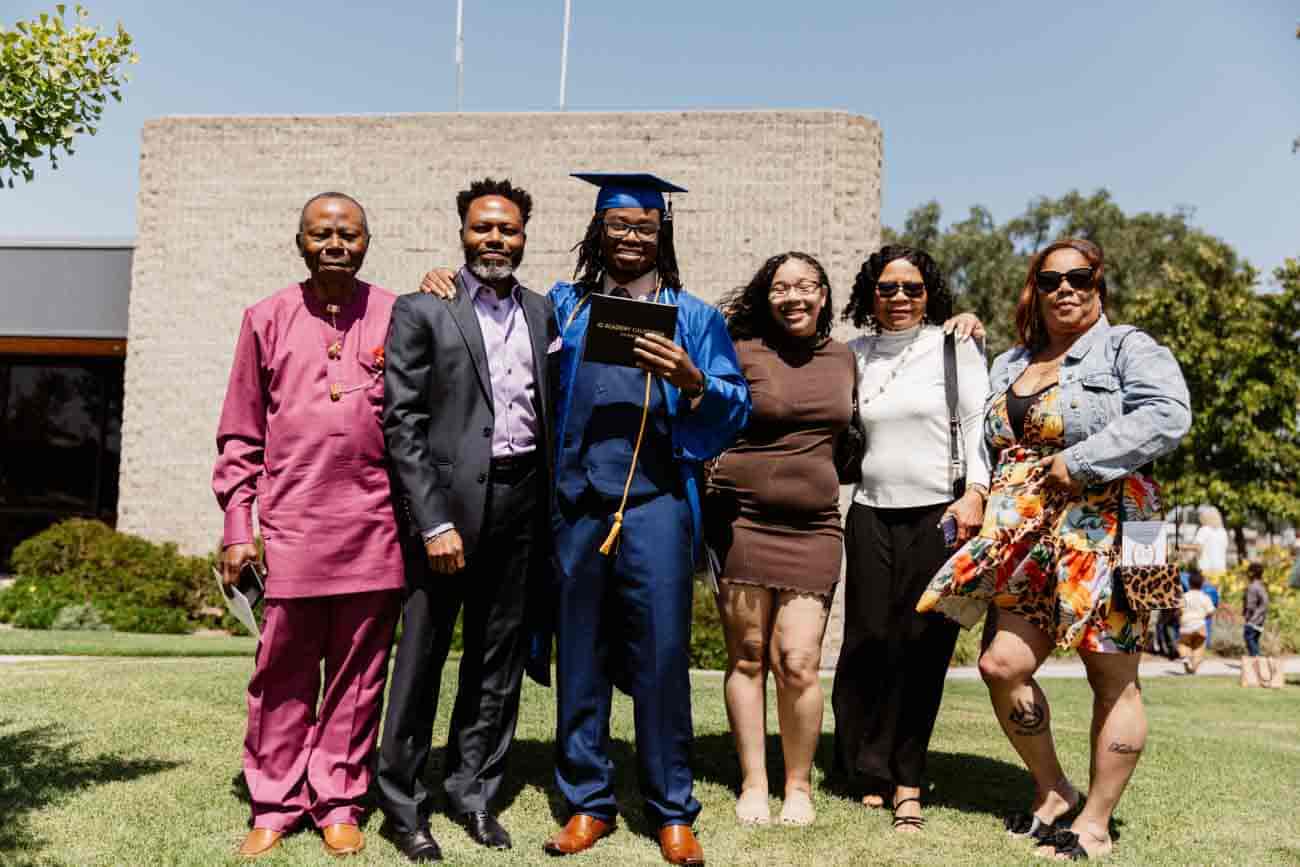 A graduate in a blue cap and gown holds a diploma, surrounded by five smiling family members on a sunny day, conveying pride and celebration.