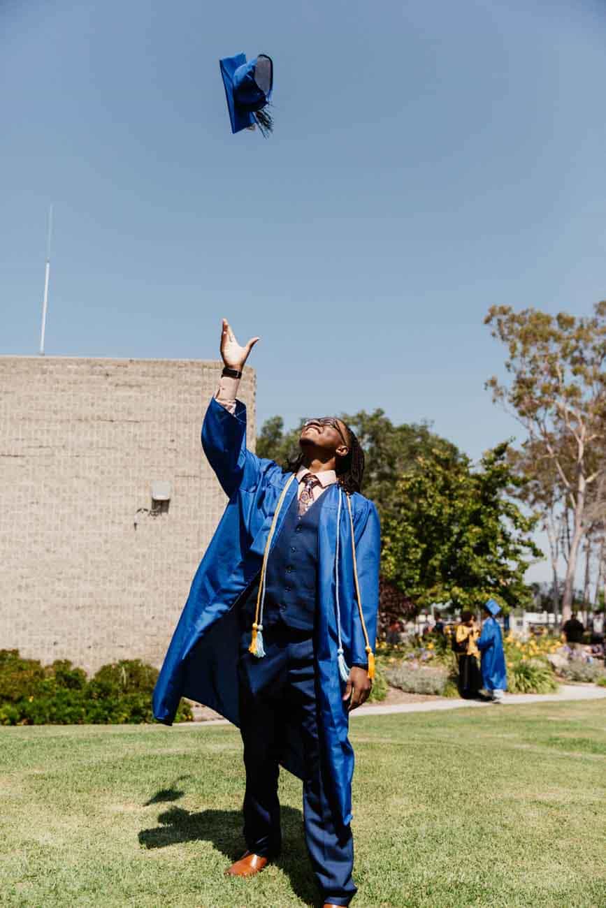 A graduate in a blue cap and gown joyfully tosses their cap into the air on a sunny day, standing on a green lawn with trees and a building in the background.