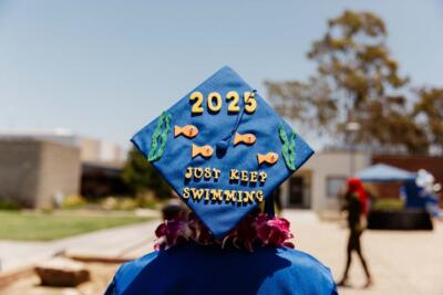 A graduate in a blue gown wears a decorated mortarboard with "2025" and "Just Keep Swimming" with fish and seaweed. Bright, celebratory atmosphere.